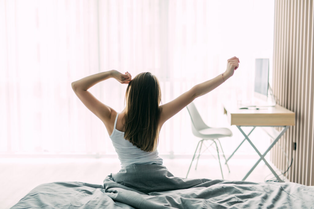 back view of young woman stretching on unmade bed after waking up and looking at city view in the window. female model relaxing at home in the morning. motivation concept