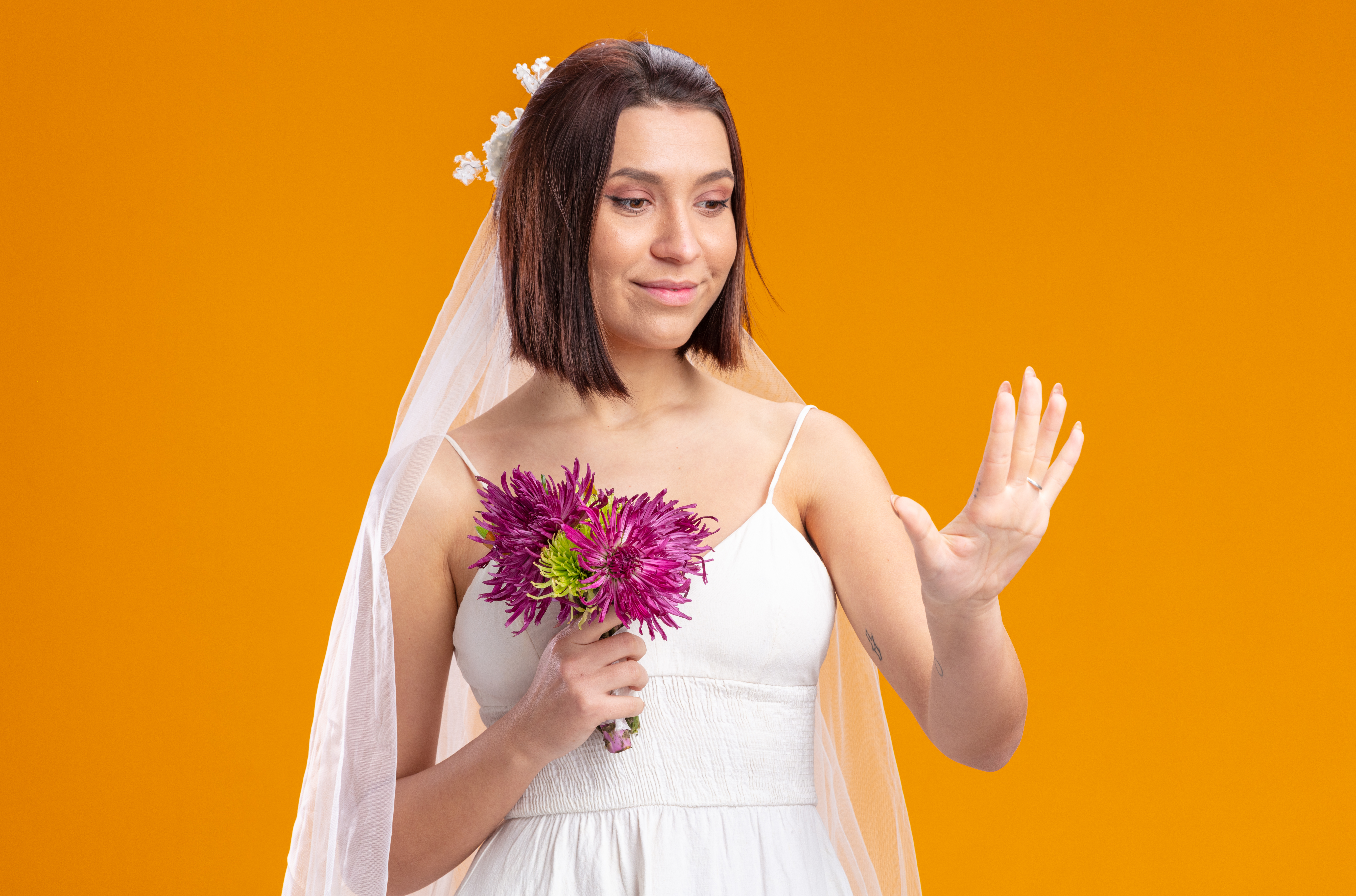 bride in beautiful wedding dress with wedding bouquet of flower looking at her ring on her finger standing over orange background