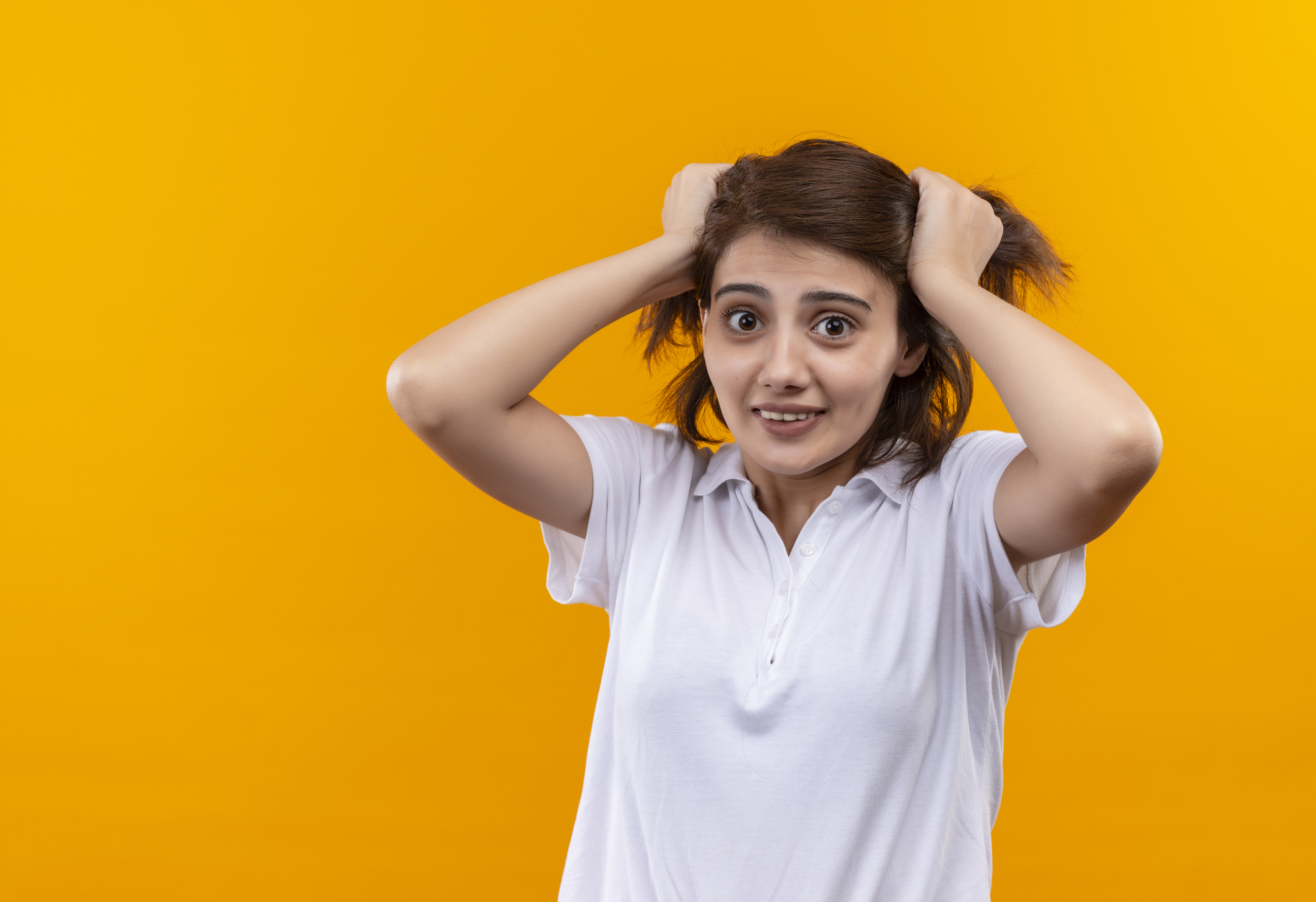 frustrated young girl with short hair wearing white polo shirt pulling her hair standing over orange background