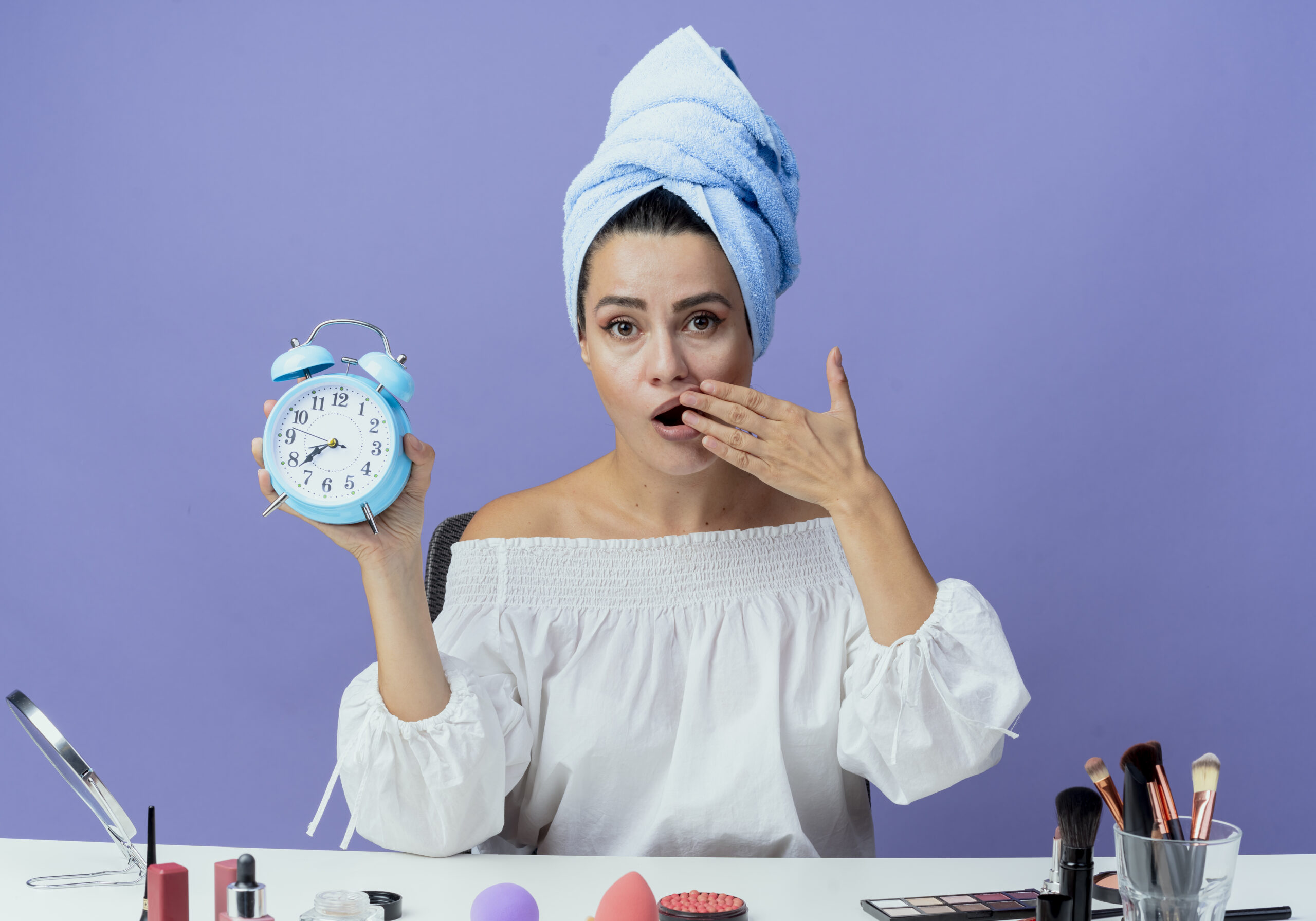 shocked beautiful girl wrapped hair towel sits table with makeup tools holding alarm clock puts hand mouth looking isolated purple wall