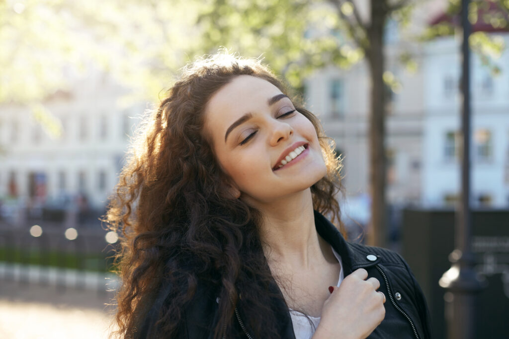lifestyle portrait of amazing young woman with genuine smile and gentle features closing her eyes in pleasure and enjoyment, feeling happy and relaxed, basking in the sunshine of spring morning